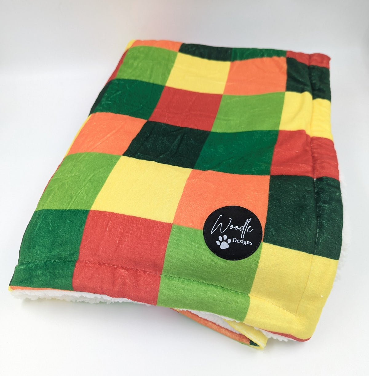 A folded checkered blanket with a plush sherpa lining, featuring a colorful patchwork pattern of green, yellow, orange, and red squares. A black circular 'Woodle Designs' logo patch is sewn onto the front. plush pet dog planket. Ireland and worldwide.