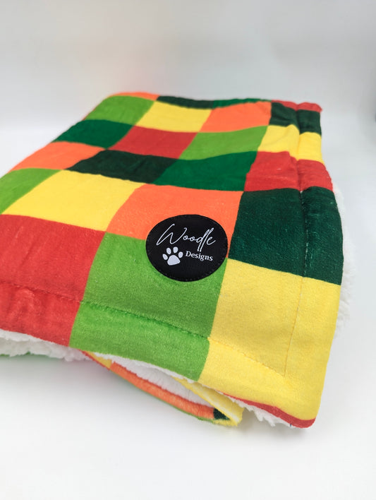 A folded plush pet blanket featuring a colorful patchwork design of green, yellow, orange, and red squares, with a white sherpa lining visible along the edge. A black circular patch on the blanket displays the brand name 'Woodle Designs'. Ireland and worldwide fast shipping
