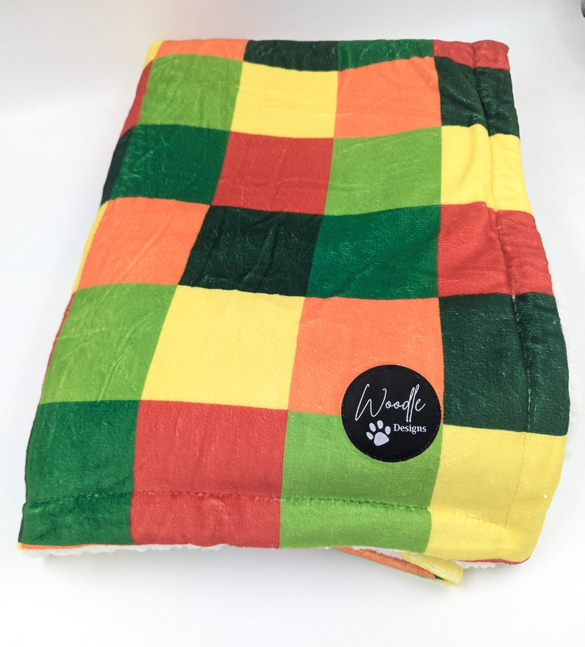 A square quilted pet dog blanket with a colorful patchwork checkered pattern of green, yellow, orange, and red, featuring a black circular 'Woodle Designs' brand patch on the front. Ireland and worldwide