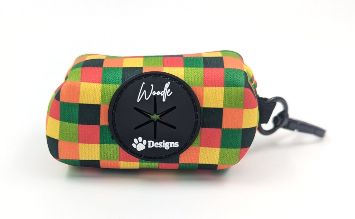 A small zippered poo bag holder for dogs with a colorful checkered pattern in green, yellow, orange, and black. The front features a black circular patch with the text 'Woodle Designs' and a paw print logo, with a black zipper pull attached on the right side. ireland wand worldwide. fast shipping